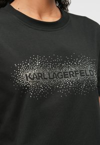 Melnas kokvilnas T-krekls ar rhinestone logo, kurā rakstīts "KARL LAGERFELD", ar teksturētu, spīdīgu rakstu uz krūtīm.
