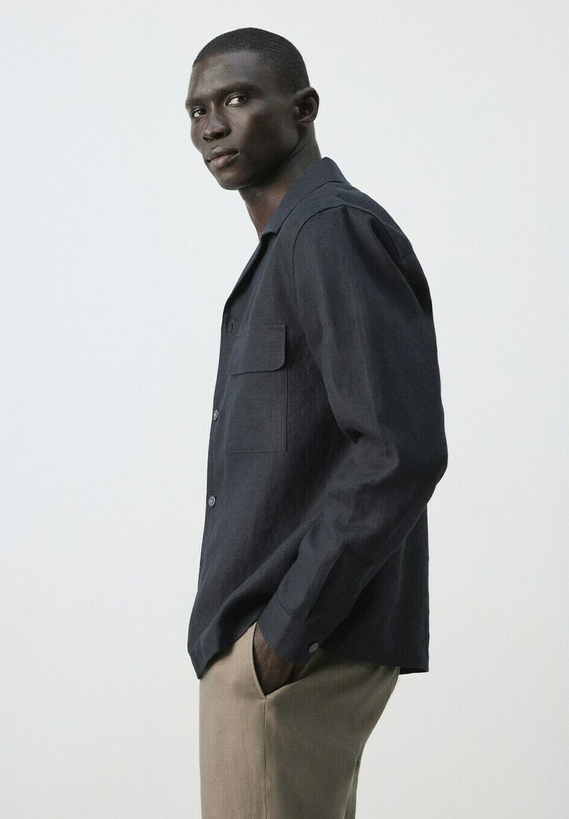 Homme portant une chemise boutonnée sombre et un pantalon beige clair, debout de profil avec une main dans la poche, devant un fond clair uni.