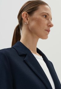 Marineblazer aus strukturiertem Stoff, mit scharfen Revers, getragen über einem weißen Hemd. Dezent sichtbare Creolen. Profilansicht.