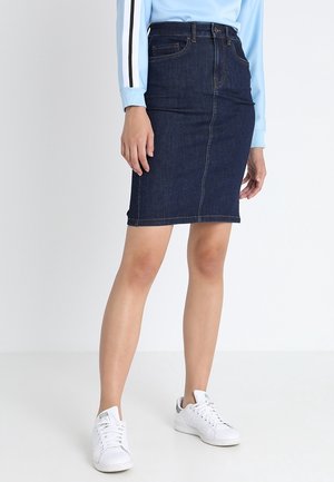 Personne portant une jupe en jean bleu foncé, un haut à manches longues bleu clair avec des rayures noires et blanches, et des baskets blanches, debout devant un fond blanc.