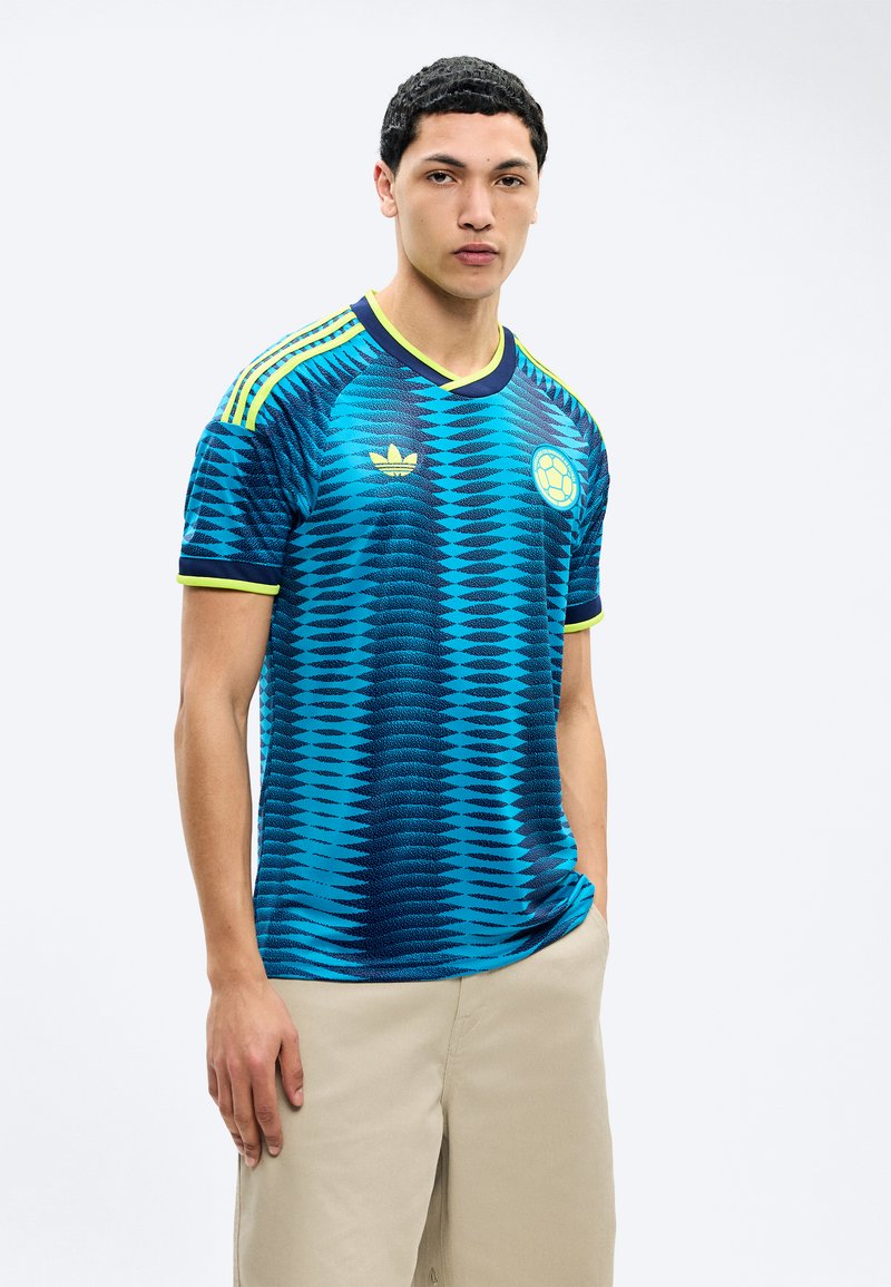 Jeune homme portant un maillot de football bleu et marine à motifs avec des accents jaunes et un pantalon beige, debout avec une main dans la poche sur fond blanc.