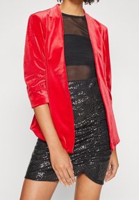 Femme portant une veste rouge en velours ouverte sur le devant, un haut noir transparent et une mini-jupe noire à sequins, debout avec un bras détendu le long du corps.