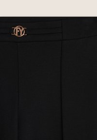 Pantaloni neri su misura con una texture liscia, caratterizzati da pieghe frontali e un dettaglio metallico circolare in tono oro alla vita.