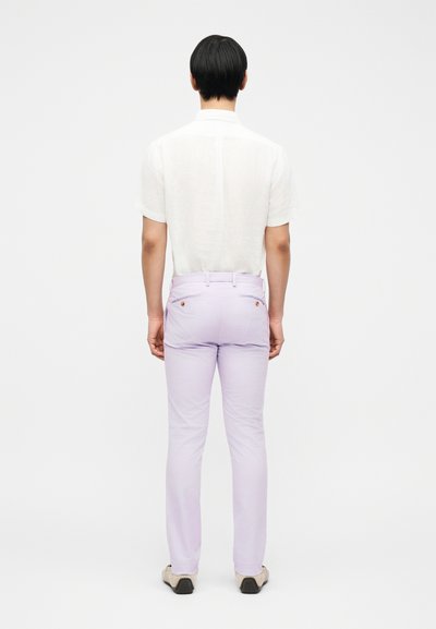 Άτομο με κοντά μαύρα μαλλιά που φοράει λευκό κοντομάνικο πουκάμισο, ανοιχτό μωβ παντελόνι και μπεζ παπούτσια, στέκεται με το πρόσωπο προς ένα απλό λευκό φόντο.