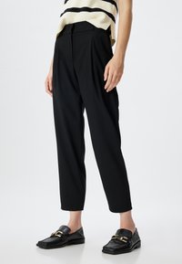Sorte tapered bukser med plisseret front og bæltestropper, lavet af et glat stof. Parret med teksturerede sorte loafers med gyldent hardware.