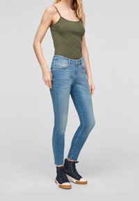Groene mouwloze top, blauwe skinny jeans met vervaging, en marineblauwe hoge sneakers met witte zolen. Gekleed voor een casual, moderne uitstraling.