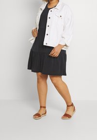 Veste en denim blanc avec des boutons marron, portée sur une robe noire jusqu'aux genoux avec un ourlet volanté, associée à des sandales compensées marron.