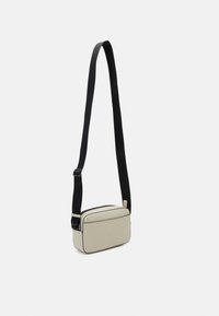 Rechthoekige schoudertas in lichtbeige materiaal met een zwarte verstelbare band, voorzien van een bovenrits en zwarte accentafwerking.