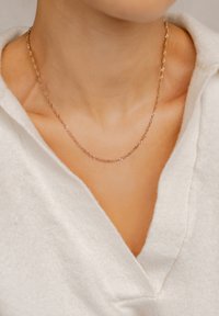 Collana in oro con un delicato design intrecciato, posata su un collo scoperto, abbinata a un morbido indumento bianco. Focus sull'estetica minimalista.