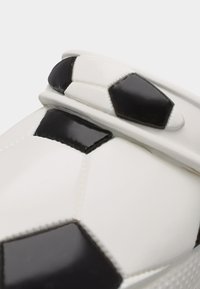 Κοντινό πλάνο ενός λευκού παπουτσιού με μοτίβο μαύρης και λευκής ποδοσφαιρικής μπάλας στο πάνω λουράκι και την περιοχή των δακτύλων.