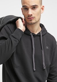 Sudadera negra hecha de tela suave, con cordones ajustables grises, un bolsillo frontal y un pequeño logotipo en parche en el pecho.