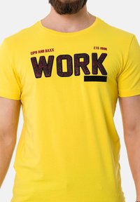 Gelbes Baumwoll-T-Shirt mit auffälligem schwarzen "WORK"-Druck, strukturierten Akzenten, Rundhalsausschnitt und kurzen Ärmeln. Markenname-Detail auf der Schulter.