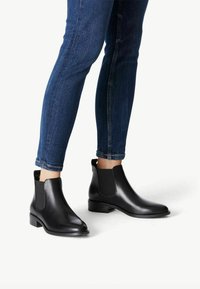 Botas Chelsea de cuero negro con paneles laterales elásticos y un tacón bajo, combinadas con jeans ajustados de mezclilla azul, que exhiben un diseño elegante.