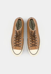 Brune canvas høyhælte sneakers med hvite gummikapsler og brune snørebånd. Svart kant langs sålen fremhever designet.