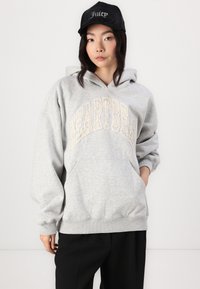 Grå hætte sweatshirt med "HEARTBEAT" trykt i cremefarve. Har en kengurulomme og ribbeløse manchetter. Bæres sammen med en sort kasket.