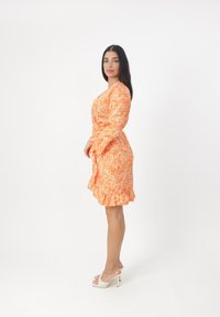 Oranges blumengemustertes Wickelkleid mit langen Ärmeln, Rüschen-Saum und bindbarem Taillendetail. Getragen mit weißen Absatzsandalen vor einem schlichten Hintergrund.
