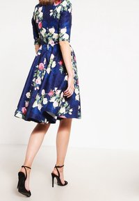 Laivansinisen kankaan kukkamekko, jossa on lyhyet hihat, pinkkejä ja valkoisia ruusuja sekä levenevä helma. Yhdistetty mustien korkokenkien kanssa.