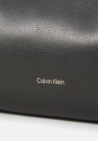 Borsa a tracolla in pelle nera caratterizzata da una texture sottile e un logo "Calvin Klein" in oro impresso sul lato. Design compatto.
