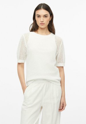 Frau mit langen dunklen Haaren, die einen weißen kurzärmeligen Strickpullover mit Netzärmeln und weiße lockere Hosen trägt, steht mit einer Hand in der Tasche.