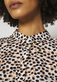 Chemise à col et manches courtes, présentant une base blanche ornée de taches abstraites noires et beiges. Fermeture à boutons avec un léger éclat et une texture lisse.