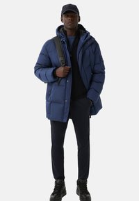 Mørkeblå pufferjakke med hætte, lynlåslommer og quiltet design. Sammen med mørke bukser og sorte snøre-sneakers.