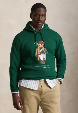 Mann iført en mørkegrønn hettegenser med en Polo Bear-grafikk som holder en golfkølle, beige bukser og en stripete skjorte under.