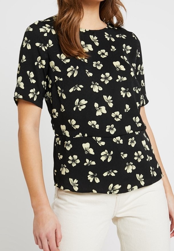 Zwarte blouse met korte mouwen en lichtgeel bloemmotief, gecombineerd met een witte broek, weergegeven van midden torso tot nek.