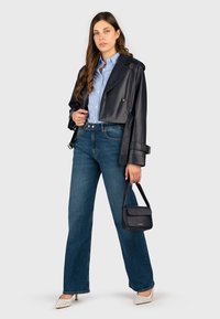 Giacca cropped in ecopelle nera, camicia a righe blu, jeans a gamba larga in denim, tacchi bianchi con borchie e una piccola borsa nera.