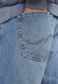 Helle blaue Jeans aus Denim mit einer خلفtasche, die ein gewelltes Nähmuster aufweist, strukturierten Stoff und kontrastierende braune Nähte.