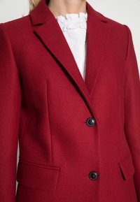 Blazer rojo a medida con un corte estructurado, solapas con muesca, dos bolsillos frontales y cierre de doble botón; fabricado en un tejido texturizado.