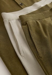 Două perechi de pantaloni tricotate, una verde măsliniu și una bej, pliate pentru a arăta talie elastică și buzunare laterale pe un fundal de material verde măsliniu asortat.