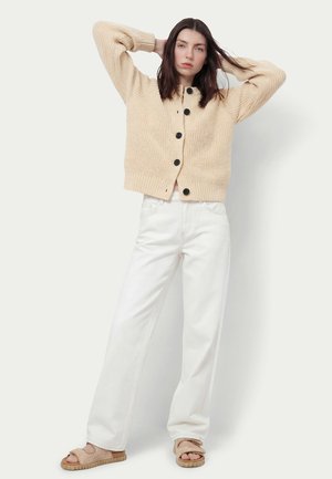 Donna con capelli scuri che indossa un cardigan beige a maglia con bottoni, jeans bianchi a gamba larga e sandali beige, in piedi con le mani sulla testa.