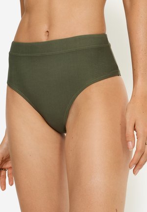 Frau trägt olivgrüne Bikinihose mit hohem Bund, gezeigt vom Bauch bis zum oberen Oberschenkel, mit entspannt an den Seiten hängenden Händen.