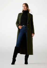 Palton lung din lână de culoare verde măsliniu, cu guler decupat, purtat peste un pulover negru cu guler înalt și blugi albaștri. Modelul poartă cizme negre cu toc.