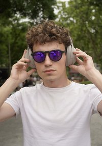 Lekkie, nauszne słuchawki w jasnoszarym kolorze. Okrągłe czarne okulary przeciwsłoneczne z lustrzanymi fioletowymi soczewkami. Osoba nosząca ma kręcone włosy i prostą białą koszulkę.