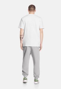 Mężczyzna z tatuażami w zwykłej białej koszulce, jasnoszarych spodniach dresowych i białych trampkach ze zielonymi detalami, stojący tyłem.