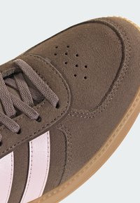 Smeđe velurove tenisice s ružičastim kožnim prugama, okruglim vrhom, perforacijama na gornjem dijelu i potplatom od gumene gume. Ravne vezice i detalji šavova.