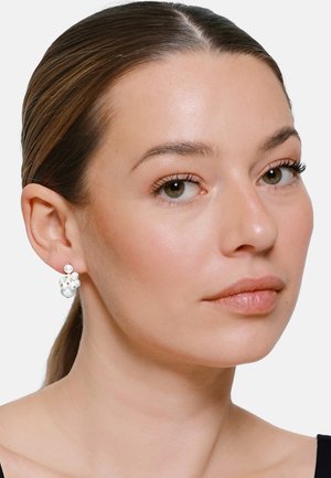 Frau mit heller Haut und braunen Haaren trägt Cluster-Perlenohrringe und ein schwarzes Oberteil, blickt leicht nach rechts mit neutralem Gesichtsausdruck.