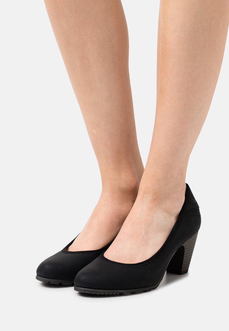 Pieds chaussés de talons hauts noirs à bouts fermés avec talons blocs, montrant une peau lisse et un fond neutre.