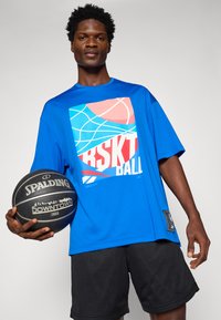 Liela zila T-krekls ar grafisku dizainu, kurā redzami basketbola motīvi, valkāts ar melniem tīkla šortiem. Melns basketbols rokās ar Spalding zīmolu.