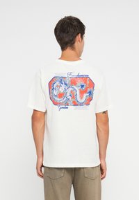 White cotton t-shirt featuring a blue and red dragon graphic on the back, hexagonal outline, and text detailing. Short sleeves, loose fit.