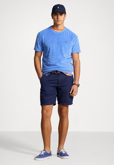 Tricou albastru din bumbac frotir cu guler rotund, pantaloni scurți navy și pantofi din pânză albaștri, completat de o șapcă închisă la culoare cu un logo.