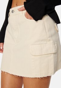 Gonna mini in denim beige con due tasche laterali, orlo sfrangiato e chiusura frontale con bottoni, indossata da una modella che porta un top nero.