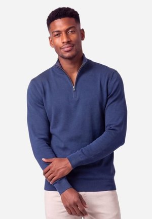 Un homme pose devant un fond uni et clair, vêtu d’un pull bleu marine à fermeture zippée courte et d’un pantalon clair.