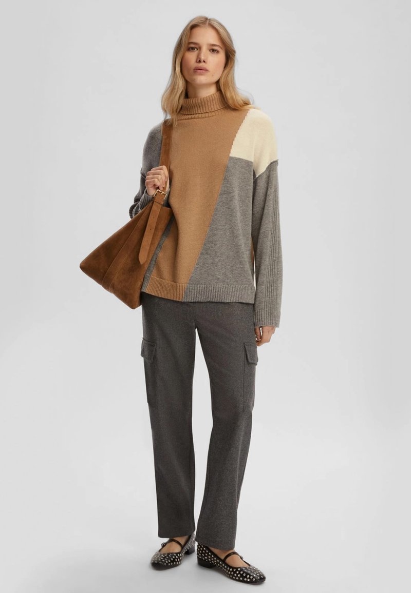 Maglione oversize marrone e grigio a motivo con colletto alto, pantaloni cargo grigi e ballerine bianche e nere a pois. Borsa in suede marrone.
