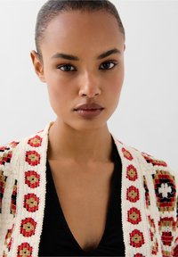Geweven cardigan met een crèmekleurige basis, voorzien van rode en zwarte bloemmotieven. Model draagt een zwarte top. Gladde textuur en open ontwerp.