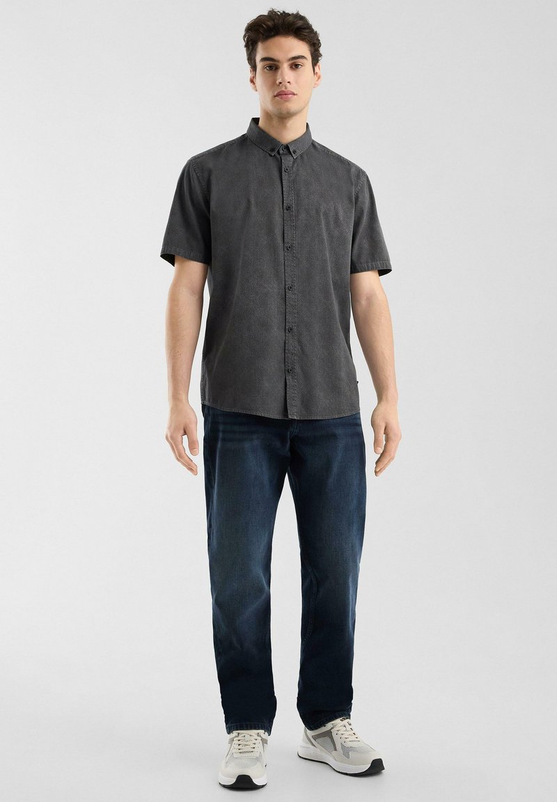 Camisa de botones de manga corta en color carbón con un patrón texturizado, combinada con jeans azul oscuro y zapatillas blancas.