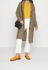 Brun texturerad lång kappa, gul skjorta med knappar, vita vida byxor och orange slip-on skor. Tillbehör inkluderar en svart handväska.