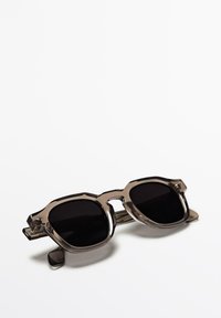 Occhiali da sole con una montatura spessa e trasparente di colore marrone e lenti scure. Presentano forme geometriche e una leggera lucentezza sulla superficie.