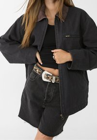 Black canvas jacket with a corduroy collar, front zipper, and large pockets. Worn over a black top, paired with black shorts and a leopard print belt.